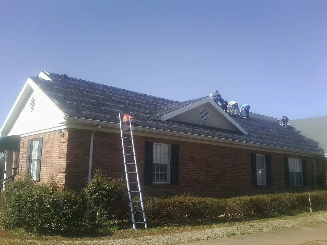 Crew tearing shingles off a brick ranch roof with a ladder set — Emergency Roof Repair work by our crew in West Bloomfield