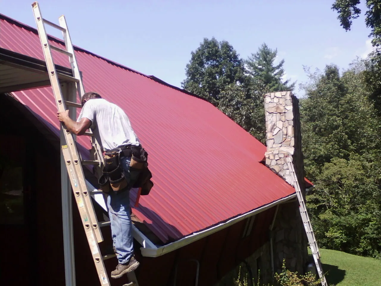 Roofer on a ladder beside a red metal roof and stone chimney — Soffit & Fascia Repair by Roofing Brothers in West Bloomfield